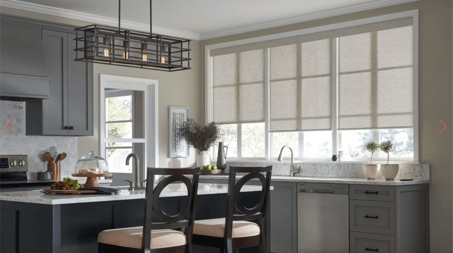 Modern kitchen with gray cabinets, marble countertops, and a large window with Roman shades over the sink area.