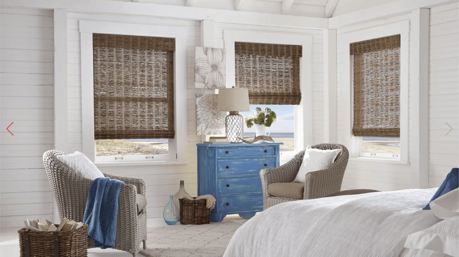 Coastal bedroom with white shiplap walls, wicker chairs, a blue distressed dresser, and woven shades by beach windows.