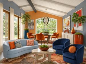 Open living-dining room with an orange accent wall, vaulted white ceiling, and a large window letting in natural light around an orange-dined table with six chairs; contemporary decor.