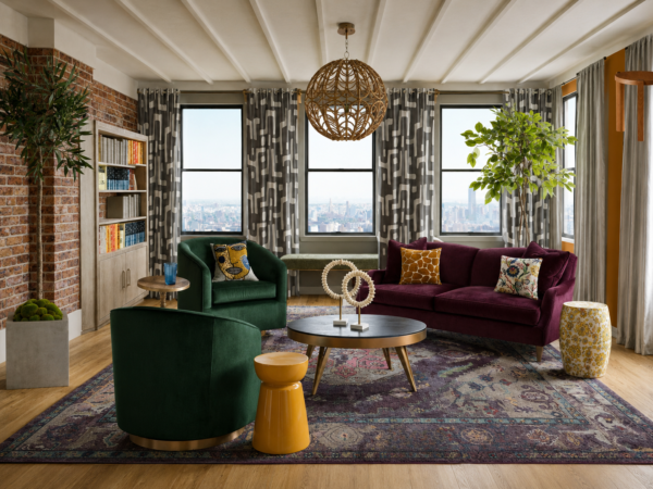 Contemporary living room with a brick wall, large windows, and green velvet chairs facing a purple sofa under a woven pendant light.