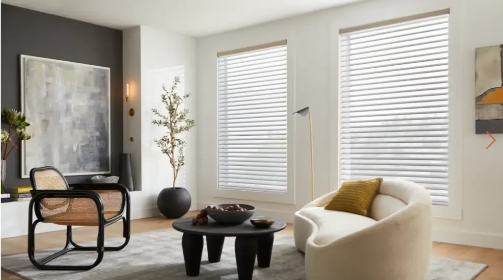 Modern living room with white walls, large windows with blinds, a cream curved sofa with a mustard pillow, a black round coffee table, and a woven-armchair beside an abstract wall art piece.