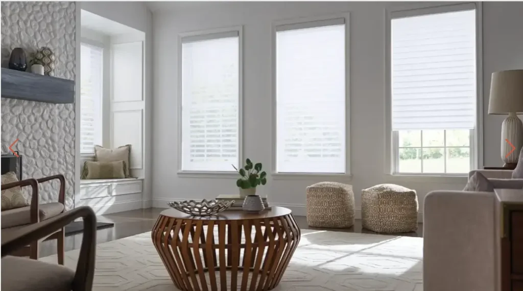 Bright living room with three large windows, woven coffee table, and neutral seating including a window nook and two patterned poufs.
