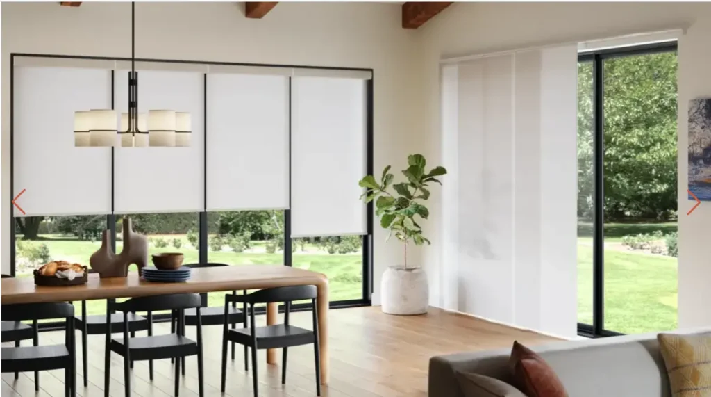 Bright open dining area with a wooden table, black chairs, a modern chandelier, and white roller blinds by large windows overlooking a green lawn.