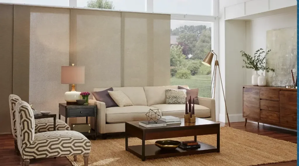Modern living room with a beige sofa, two patterned chairs, a dark wood coffee table, and a woven rug under natural light from large windows.
