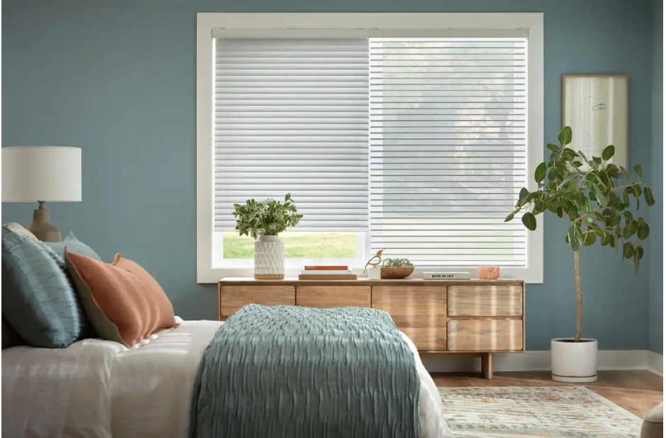 Bedroom with blue walls, a wooden dresser under a large window with white blinds, and a bed with teal and peach cushions plus a potted plant on the dresser.