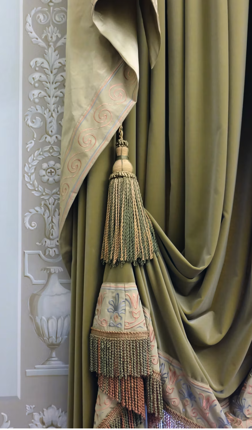 Close-up of an olive-green, heavy drapery with gold and pale-embroidered tassel fringe against a decorative white wall panel and vase motif.