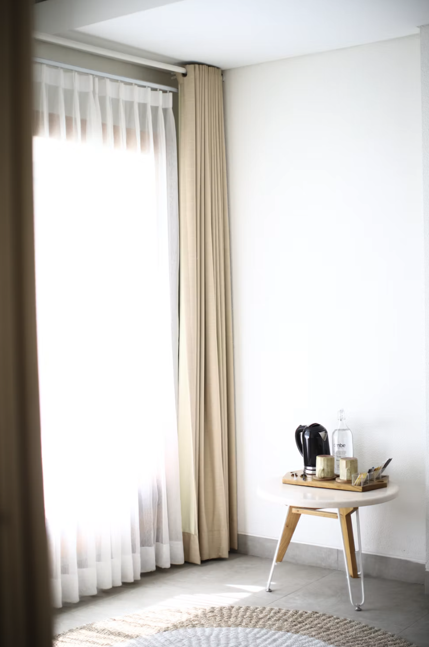 Bright corner of a minimalist room with sheer white curtains, beige drapes, and a round table holding a kettle, cups, and a bottle.